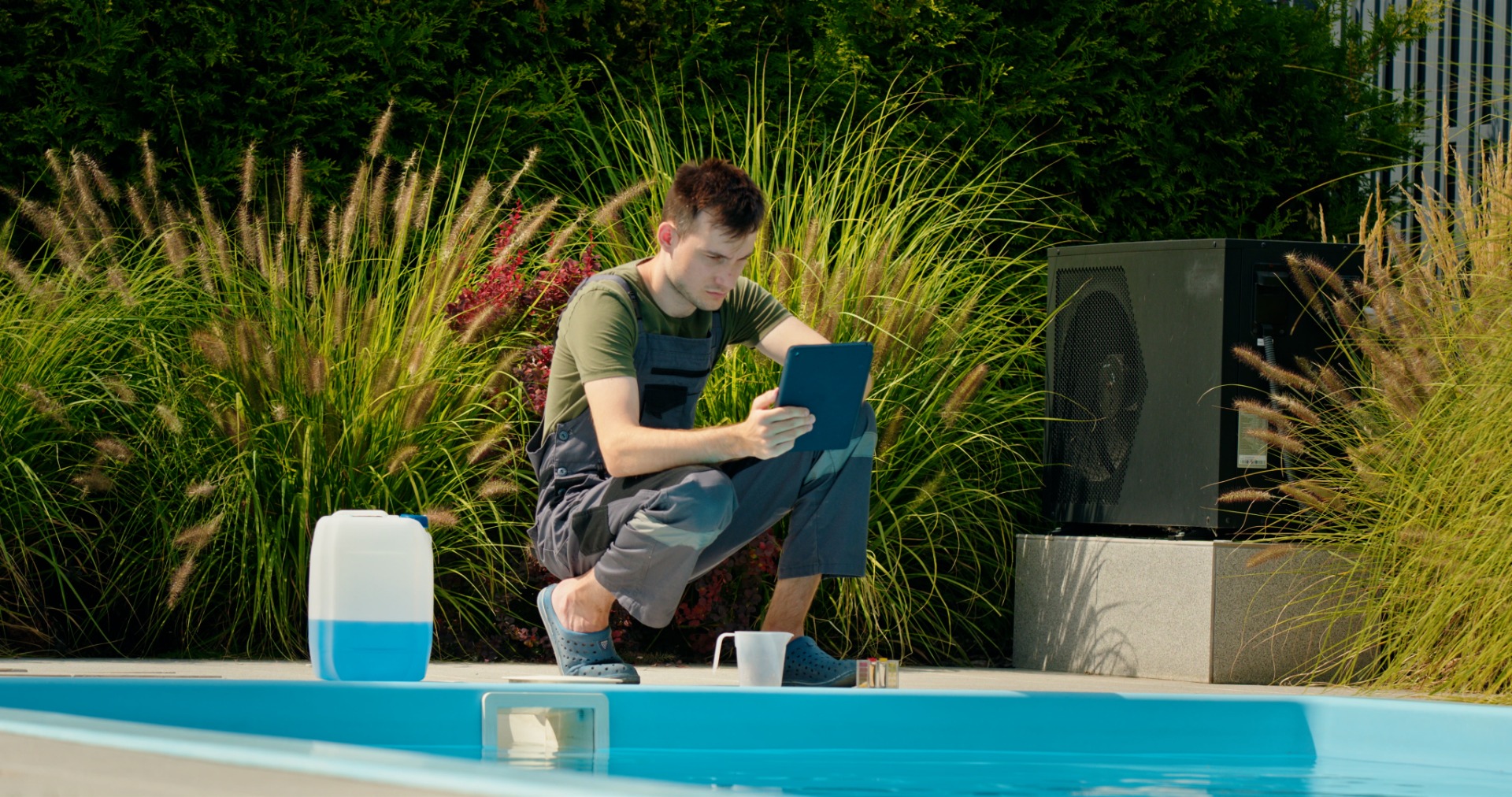 pool-technician-using-tablet-by-pool-surrounded-by-greenery-chemical-container-focused